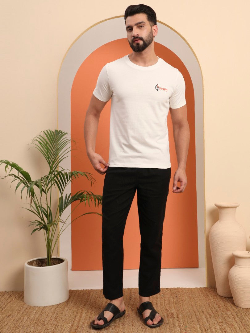 A man standing in an indoor setting wearing a white crew neck t-shirt with the text 'OM TAT SAT' on the front and black pants. He is also wearing black sandals and the room has a plant to his left and a beige chair in the background.