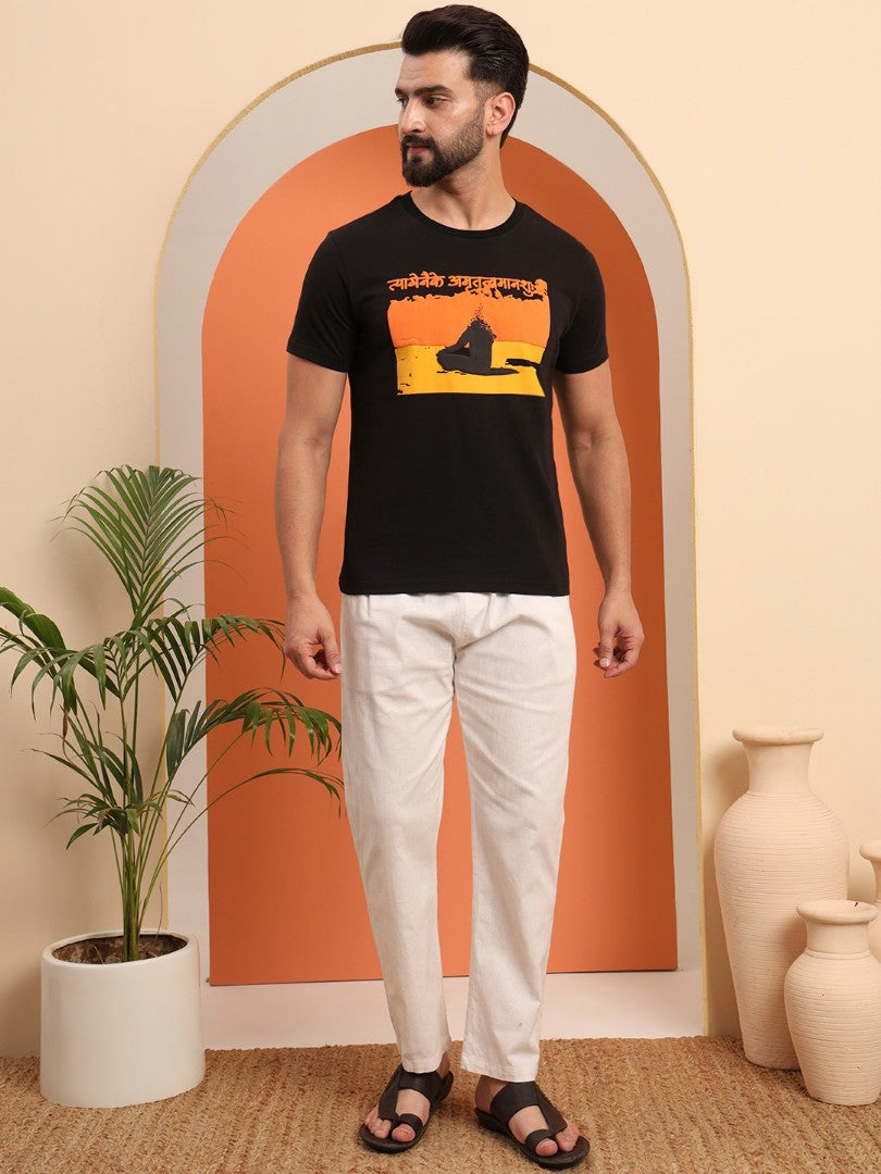 A man standing in an archway wearing a black t-shirt with a colorful graphic and text, paired with white pants and black sandals.